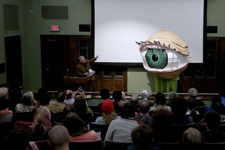 A professor lecturing to a class with a large eyeball sculpture beside him