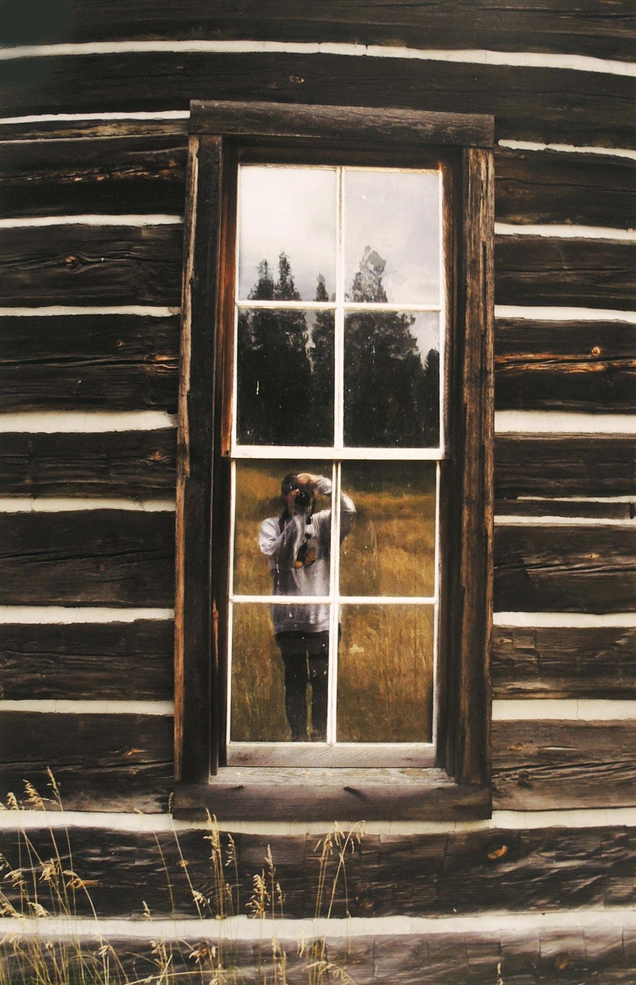 A photograph by Gabrielle Robinson of the photographer mirrored in the window of a log cabin