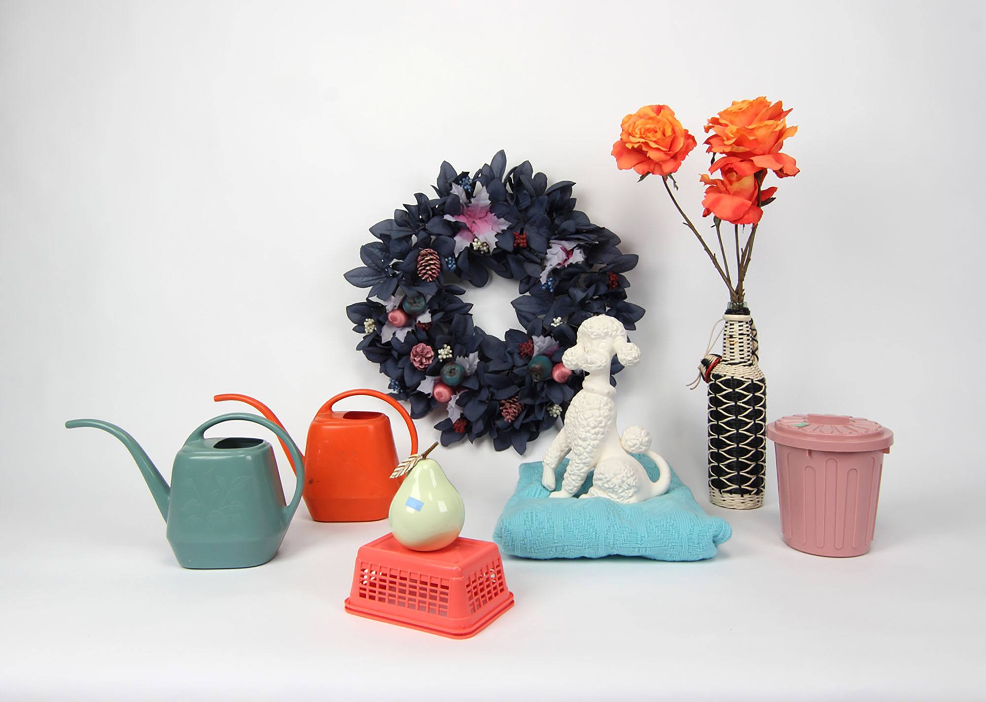 A still life of watering cans, a trash can, a poodle sculpture, a wreath, a pear sculpture, and flowers