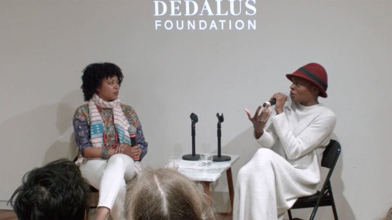 Two individuals sitting in front of a projection of the Dedalus Foundation logo, one speaking into a microphone
