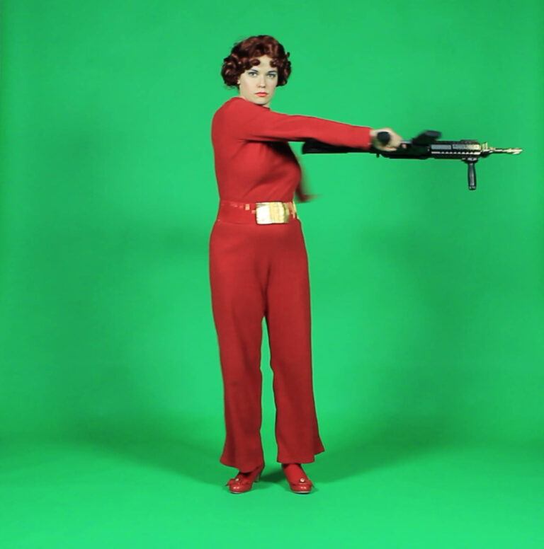 A woman dressed in a red jumpsuit carrying a gun in one hand on a green screen