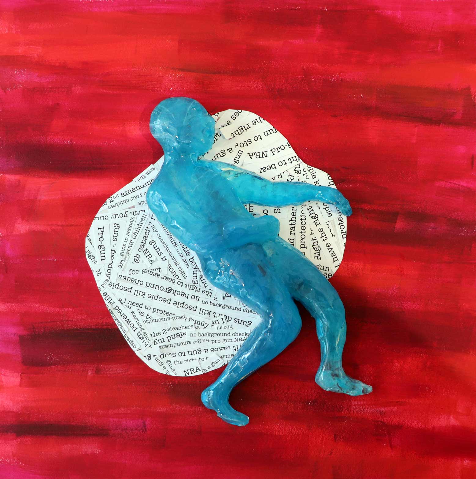 A small sculpture of a person laying down on a red sheet, with text fragments formed in the shape of a pool of blood with text relating to gun violence