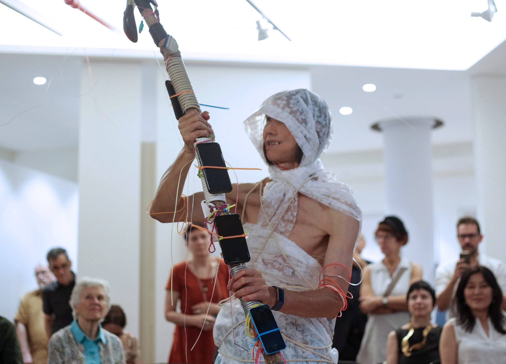 A person performing with fabric wrapped around their head and body holding a long stick with iPhones held on while people watch