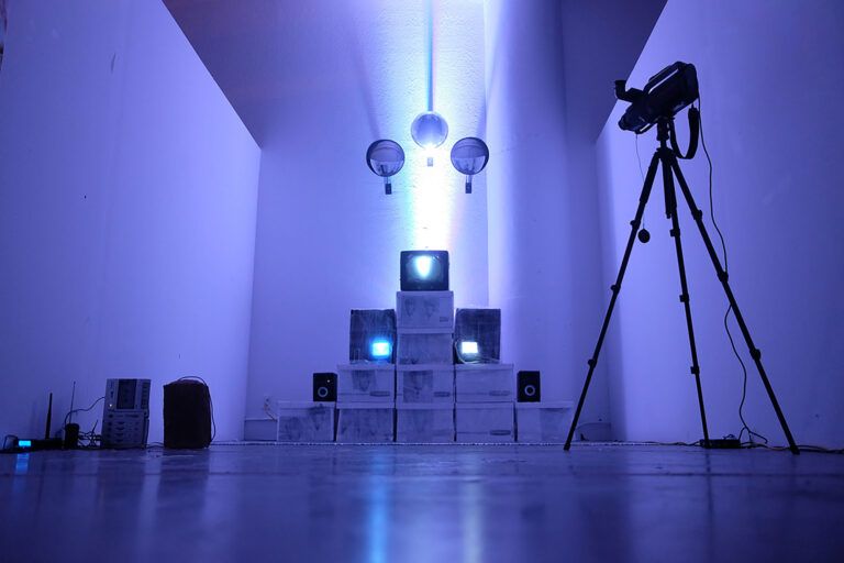 A gallery installation containing bozes, monitors, and speakers stacked in a pyramid