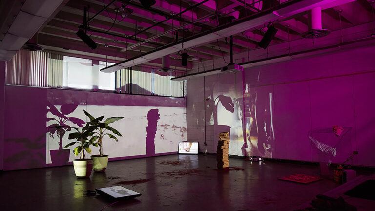A purple lit gallery space with plants and handmade structures creating shadows in wall projections