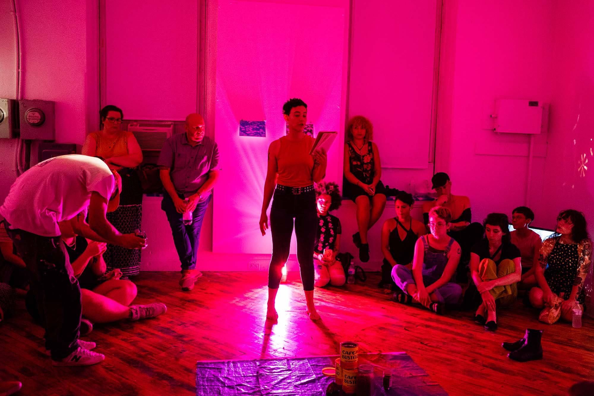 A pink lit room containing a central person reading with spectators seated around the periphery