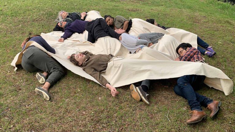 Ten people laying in the grass, connected at the waist by a canvas tarp