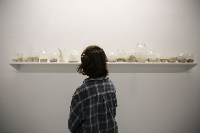 Installation view of jars on a ledge filled with clay and water. A person looks at the objects.