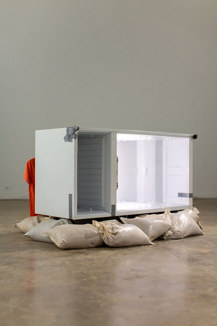 A refrigerator turned on its side atop a layer of sand bags in a gallery space