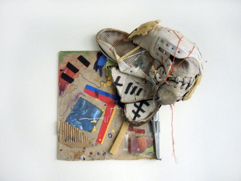 An installation image of a wall mounted artwork containing collaged papers, markings, and a baseball glove held on by string