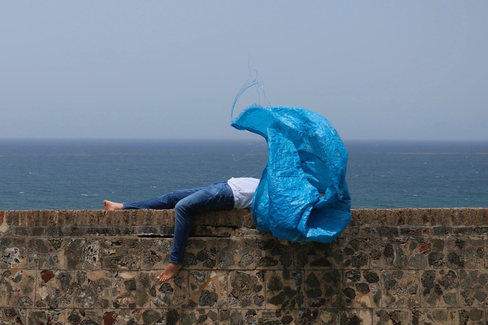 An image of a person laying on a retaining wall near the ocean, with blue plastic shading their head