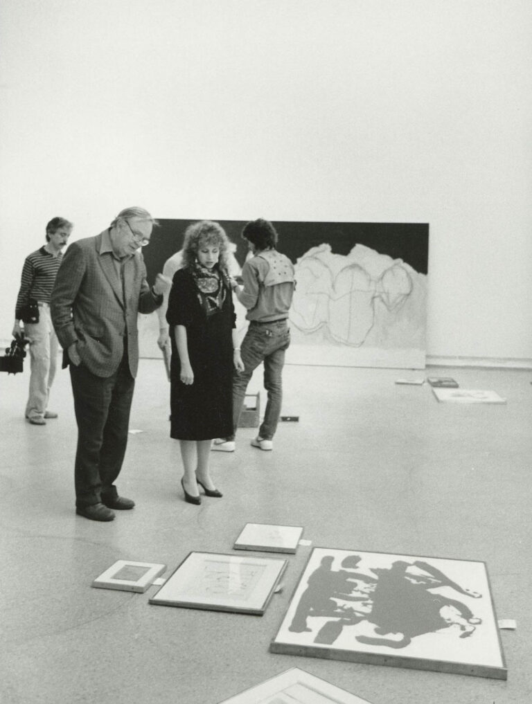 Robert Motherwell and Guggenheim Museum Director Lisa Dennison stand and look at several of Motherwell's paintings arranged on the floor of an exhibition space. Motherwell's painting "Hollow Men" rests against a wall in the background. 1984.